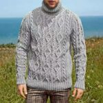 Photo d'un homme portant un pull col roulé tricoté à torsade. Derrière lui, de la pelouse et la mer.