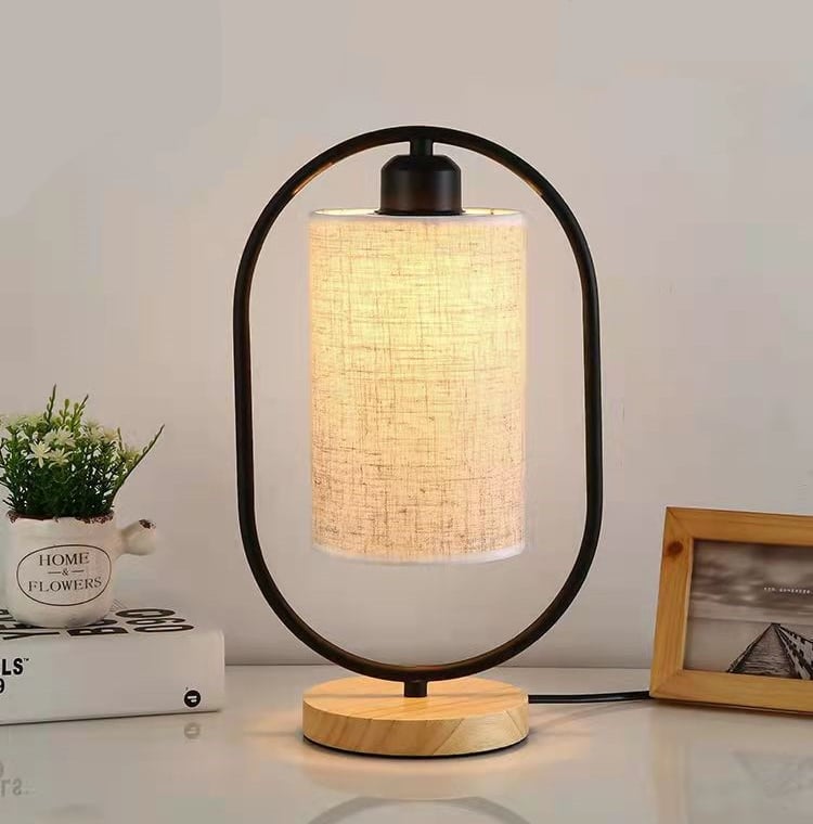 Photo d'une lampe ovale en fer noir avec un socle en bois et un abat jour cylindrique en lin. La lampe est allumé avec une faible luminosité et posé sur une table. En arrière plan, une photo et une plante.