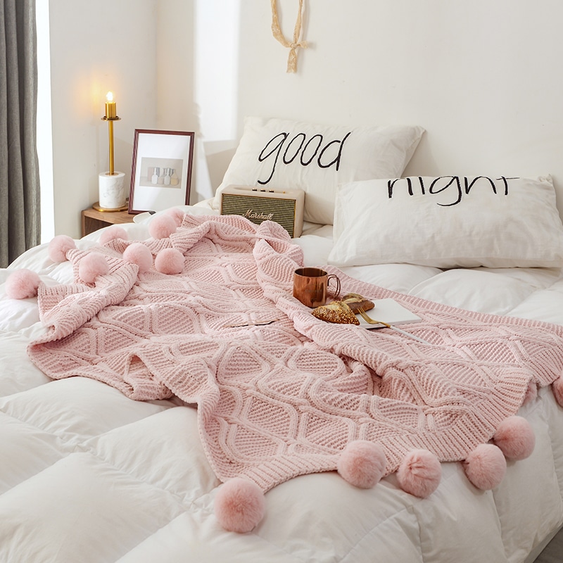 Photo d'un plait tricoté rose avec des pompons posé sur un lit aux draps blanc.