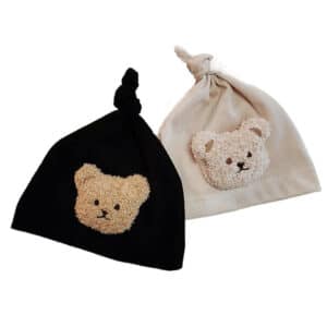 Photo de deux bonnets en coton pour bébé, un noir et un beige avec un ours en peluche dessus.