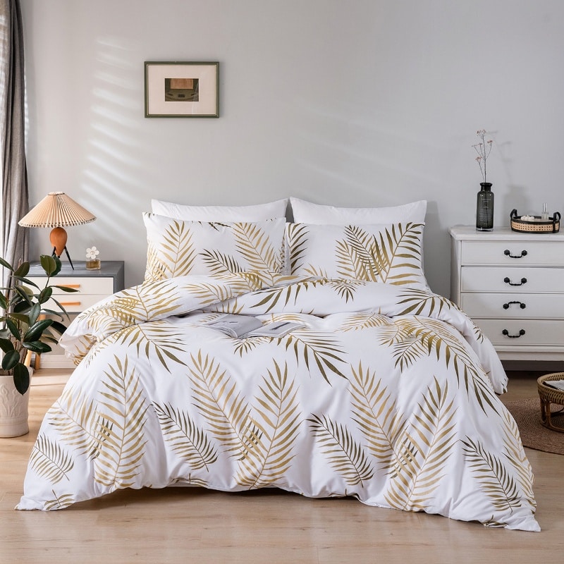 Photo d'une parure de lit blanche avec des motifs de feuilles de palier dorées dessus. La parure est sur un lit dans une chambre moderne avec du parquet, une table de nuit, une lampe, une commode et un petit cadre au mur.