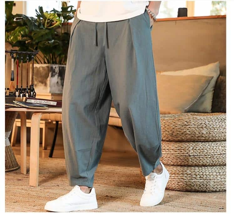 Photo d'un pantalon sarouel gris foncé avec des poches latérales, porté par un homme, les mains dans les poches avec des baskets et un tee-shirt blancs dans un décor cosy avec beaucoup de bois.
