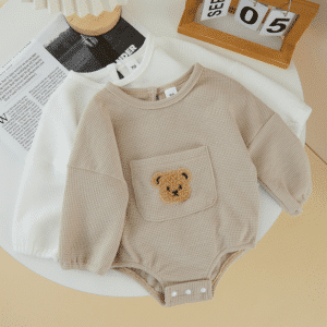 Barboteuse pour bébé à manches longue avec un ourson en peluche dessus. Une beige et une blanche sur une table en bois avec des feuilles et un calendrier moderne.