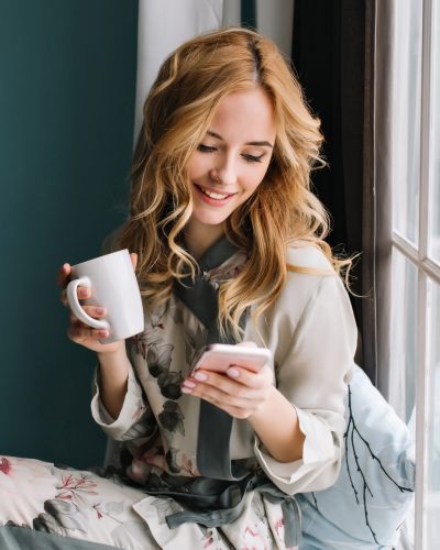 Pretty blonde girl sitting on window sill with cup of coffee, tea and smartphone in hands. She has long blonde wavy hair, smile and looking at her phone. Wearing beautiful silk pajama.